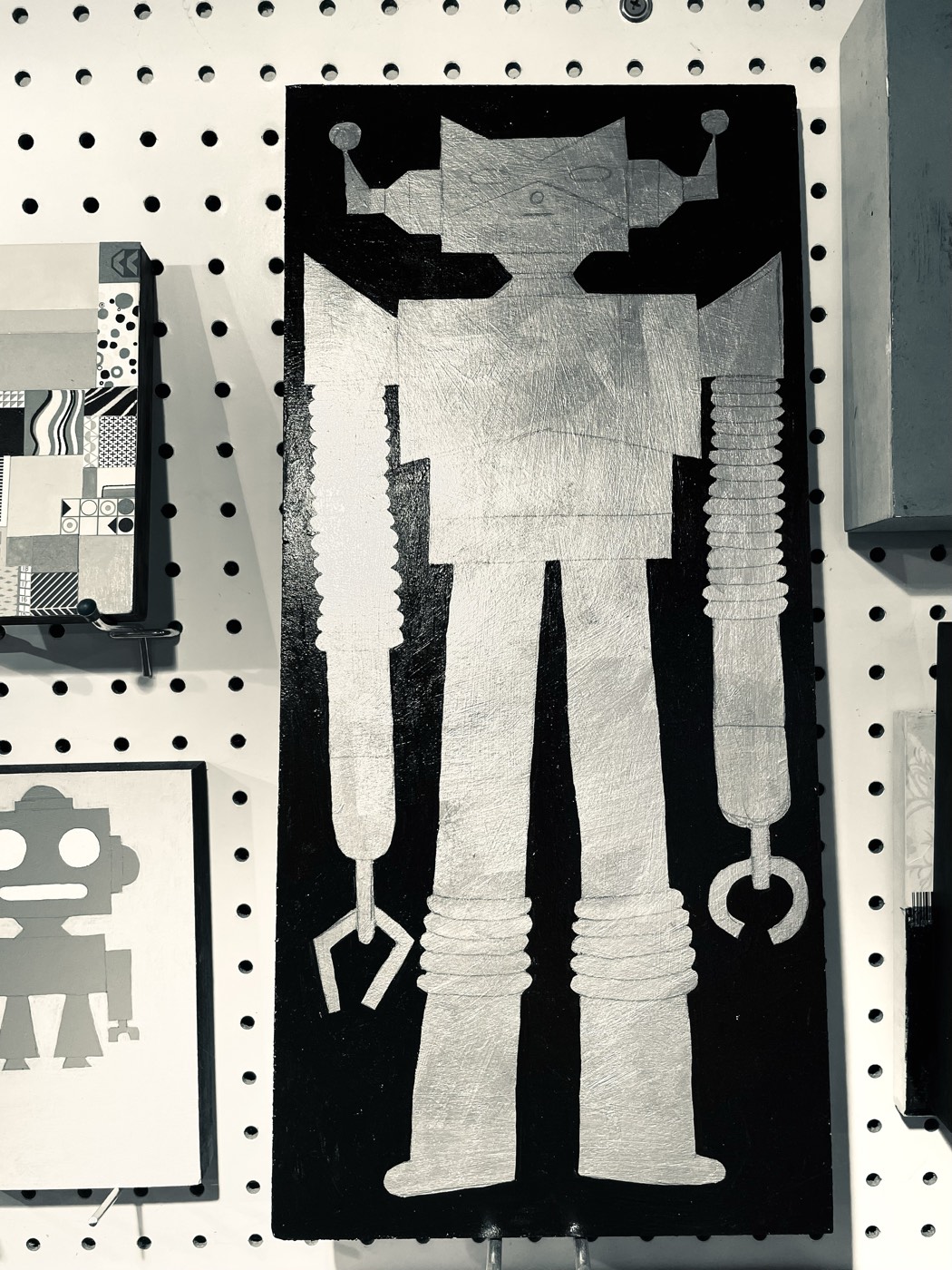 A silver robot painting by Maura Cluthe mounted on the pegboard wall